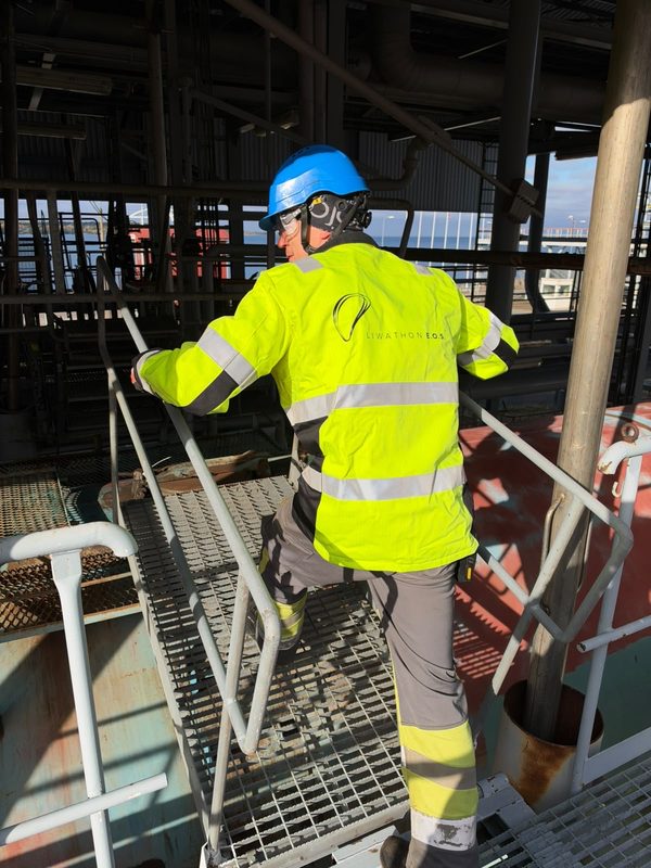 Un operario de Liwathon E.O.S. con casco se agarra a los pasamanos mientras sube la escalera de un pórtico, con el mar al fondo.