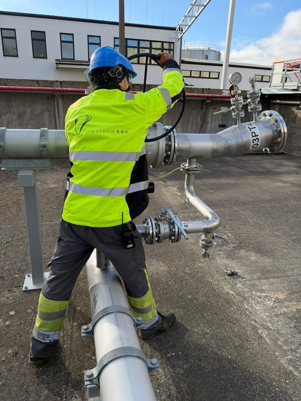 A Liwathon E.O.S. operator in a hard hat and branded high-visibility jacket turns a valve wheel on a pipeline.