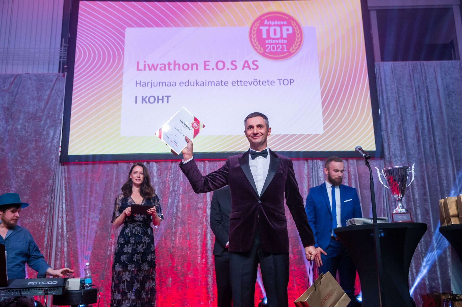 On stage at the Harjumaa TOP gala, a man in black tie raises the award; the screen behind reads “Liwathon E.O.S AS, Harjumaa edukaimate ettevõtete TOP, I KOHT” (first place).