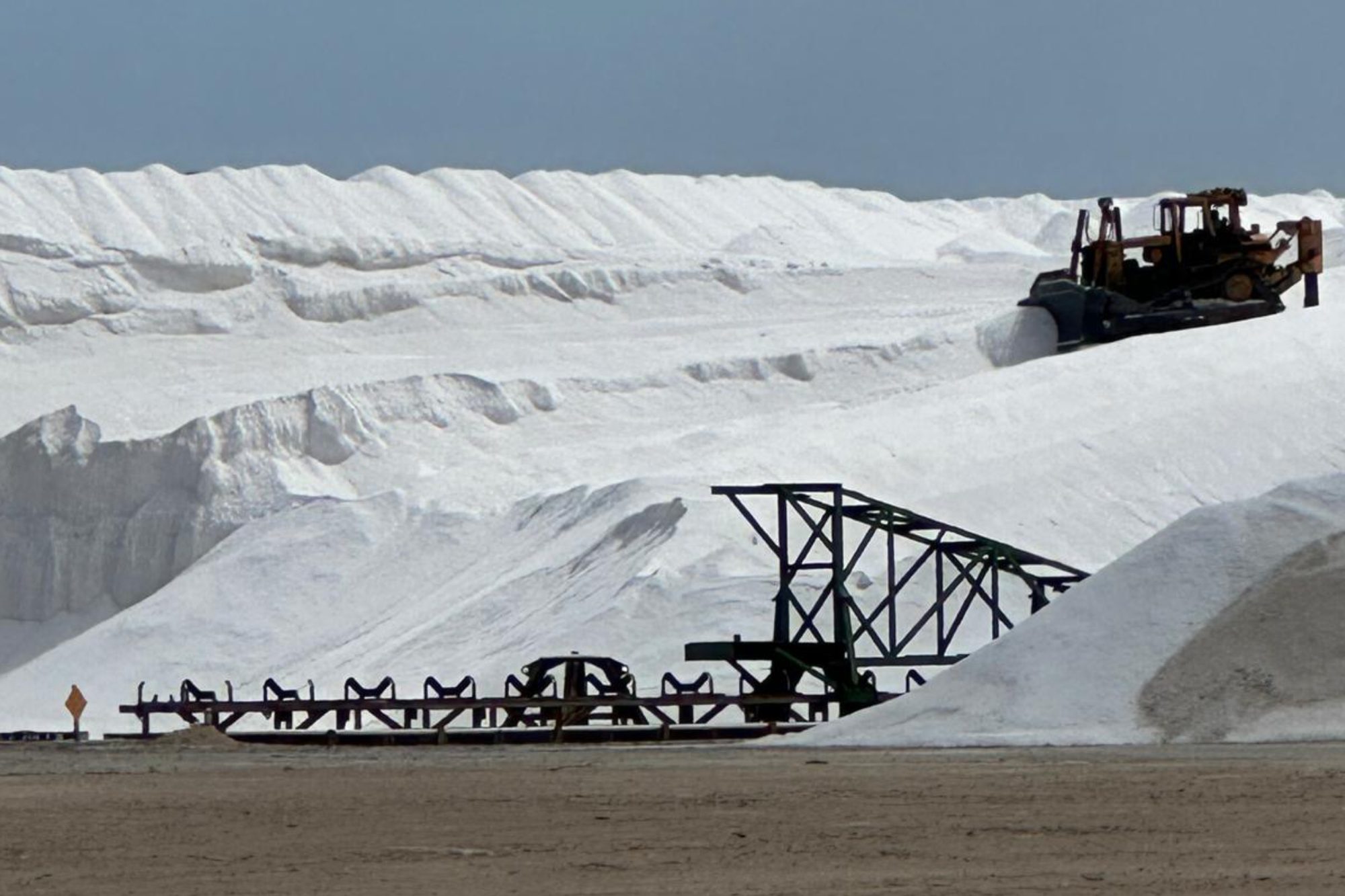 A bulldozer works high on a white salt stockpile behind the steel frame of a stacking conveyor, Great Inagua.
